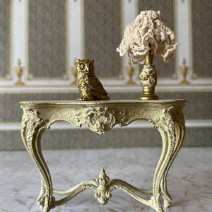 May include: A white painted, ornate, wooden table with a gold owl figurine and a lamp with a crocheted shade on top.