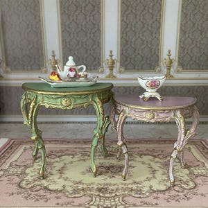 May include: Two miniature ornate side tables. One is green with gold accents, the other is pink with gold accents. A miniature tea set sits on the green table. A decorative bowl sits on the pink table. The tables have curved legs.
