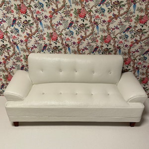 Könnte beinhalten: Ein weißes Ledersofa mit Knöpfen und einer leicht geschwungenen Rückenlehne. Das Sofa steht auf einem weißen Boden vor einer Tapete mit Blumenmuster.