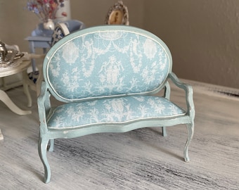 May include: A light blue miniature sofa with a white floral patterned fabric upholstery. The sofa has a curved back and scrolled legs.