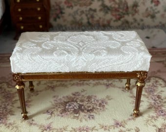 May include: Miniature upholstered bench with a white patterned top and a wooden frame. The legs are brown with gold accents. The bench is placed on a floral patterned rug, suggesting a dollhouse or miniature furniture.