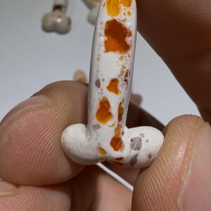 Puede incluir: Primer plano de una escultura de cerámica blanca y naranja moteada con forma de falo. La escultura se sostiene entre dos dedos. Otras esculturas similares son visibles en el fondo.