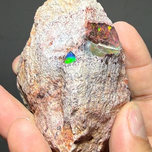 Puede incluir: Una roca rugosa beige con un ópalo vibrante y multicolor incrustado. El ópalo muestra destellos de verde, azul y rojo. La superficie de la roca tiene un aspecto texturizado, con tonos marrón rojizo y blanco. Un espécimen de piedra preciosa natural.