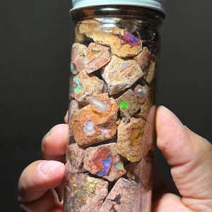 May include: A clear glass jar filled with rough-cut opal stones. The opals display a variety of colors, including blue, green, and orange, and are encased in a clear glass jar with a white lid. The jar is held against a dark background.