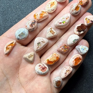Puede incluir: Una colección de piedras preciosas de ópalo en bruto de varias formas y tamaños. Los ópalos muestran una mezcla de colores, incluyendo blanco, naranja y destellos iridiscentes. Algunas piedras tienen estructuras y patrones internos visibles, con una textura rugosa y natural.