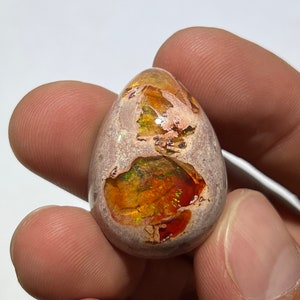 Puede incluir: Un cabujón de ópalo en forma de lágrima con una matriz marrón, blanca y verde. El ópalo muestra un arcoíris de colores, incluyendo rojo, naranja, amarillo, verde, azul y morado.