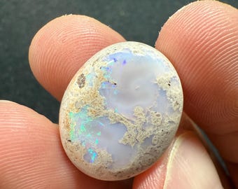Peut inclure: Une pierre d'opale ovale avec une base blanche et des éclairs irisés de bleu et de vert. La pierre a une matrice texturée, brun clair sur les bords. L'opale est tenue entre les doigts.