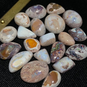 Op de afbeelding: Een verzameling van 17 natuurlijke opaal cabochons in verschillende vormen en maten. De opalen zijn een mix van wit, bruin en grijs met iriserende kleurflitsen.