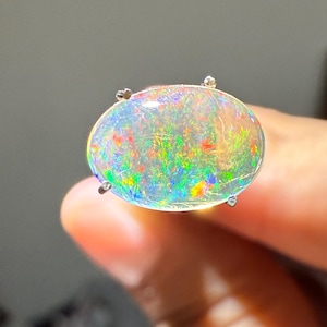 Könnte beinhalten: Ein großer, ovaler Opal-Edelstein mit einer lebendigen Darstellung von Farben, darunter Grün, Rot und Blau. Der Opal ist mit vier kleinen Zinken gefasst. Der Edelstein wird zwischen den Fingern gehalten.