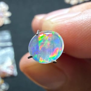 Op de afbeelding: Een ronde, doorschijnende opaal edelsteen in een zilverkleurige metalen zetting met pootjes. De steen vertoont een levendig kleurenspel met flitsen van neongroen, elektrisch blauw, helder oranje en geel over het gladde, gepolijste oppervlak.