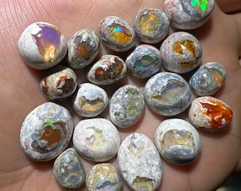 May include: A collection of 18 opal cabochons in various shades of grey, white, and brown with iridescent flashes of colour. The opals are rough and have a natural, earthy look.