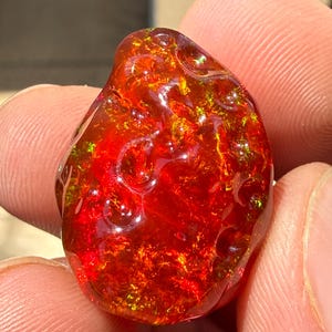 May include: A vibrant, polished gemstone with a fiery red and orange color palette. The stone has a glossy, textured surface with hints of green and yellow. The gemstone is held between two fingers, showcasing its natural beauty.