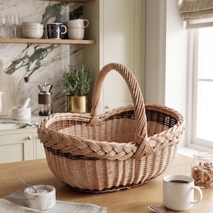 Può includere: Un cestino in vimini intrecciato marrone chiaro, con un bordo intrecciato e un manico curvo, è appoggiato su un tavolo di legno. Il cestino ovale ha un bordo marrone scuro. Una tazza bianca con caffè e una piccola ciotola di marmo sono anche sul tavolo.