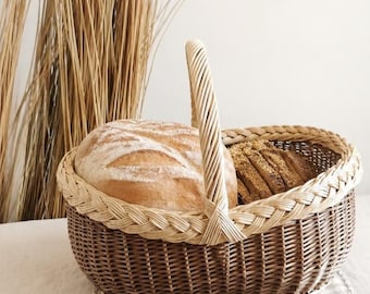 May include: A woven brown wicker basket with a braided rim and handle, filled with a large loaf of bread and sliced bread. The basket sits on a checkered cloth, with dried wheat stalks in the background.