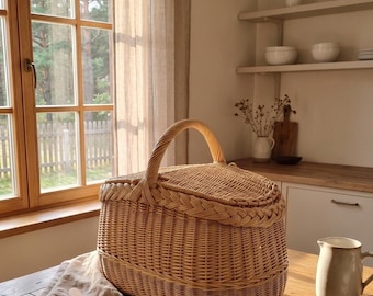 May include: A woven wicker picnic basket with a handle and a lid sits on a wooden table. The basket is light brown and has a braided trim. A beige linen napkin and a wooden butter knife are next to the basket.