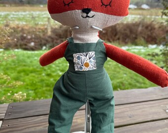 May include: A handmade plush fox toy wearing green overalls with a floral print pocket. The fox has a white face with black eyes and a black nose.