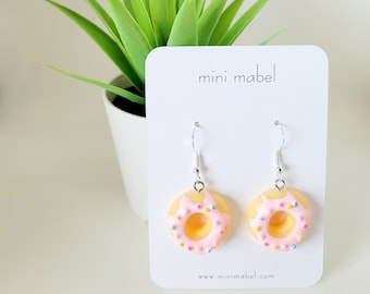 May include: A pair of donut-shaped earrings with pink icing and colourful sprinkles. The earrings are silver and have a hook fastening. The earrings are displayed on a white card with the text "mini mabel" and "www.minimabel.com".