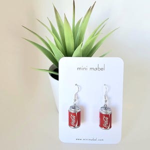 Può includere: Un paio di orecchini a forma di lattina di Coca-Cola. Gli orecchini sono repliche in miniatura dell'iconica lattina di soda, con il logo rosso e bianco della Coca-Cola. Sono appesi a ganci per orecchie color argento. Gli orecchini sono esposti su una carta bianca con la scritta "mini mabel".