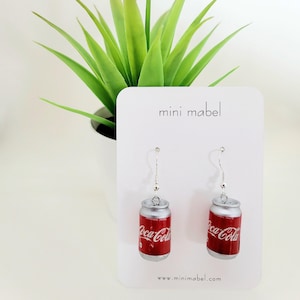 Può includere: Orecchini a forma di lattina di Coca-Cola in miniatura. Gli orecchini sono piccole repliche delle classiche lattine di soda, con il logo rosso e bianco della Coca-Cola. Sono appesi a ganci color argento. Lo sfondo include una carta bianca con il testo "mini mabel" e una pianta verde.