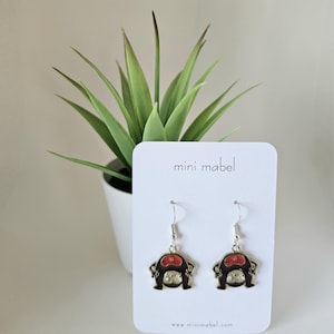 May include: A pair of silver-tone drop earrings with a cartoon-like design. Each earring features a red, brown, and silver-colored figure. The earrings are displayed on a white card with the text "mini mabel" and the website address.