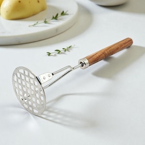 May include: A stainless steel potato masher with a wooden handle. The masher has a grid-like head for efficient mashing. The handle is made of light brown wood and is attached to the masher head with a metal connector. The masher is designed for kitchen use.