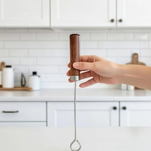 May include: A wooden-handled kitchen tool with a stainless steel shaft and a looped end. The handle is a dark brown color, and the tool is held against a white countertop. The kitchen tool is likely used for stirring or mixing.