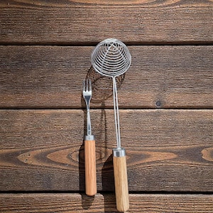 Könnte beinhalten: Eine Edelstahlgabel und ein Drahtgeflecht-Schaumlöffel, beide mit hellbraunen Holzgriffen. Die Küchenutensilien sind vor einem dunkelbraunen Holzhintergrund platziert.