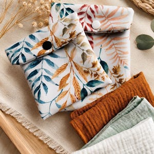 Puede incluir: Dos bolsas cuadradas de tela con fondo blanco y un estampado botánico de hojas azules, naranjas y verdes. Cada bolsa tiene una solapa plegable asegurada con un botón a presión. Junto a las bolsas hay tres paños de muselina doblados en color óxido, verde salvia y blanco.