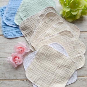 Può includere: Una collezione di morbidi asciugamani quadrati per bambini in vari colori pastello, tra cui blu, verde e beige. Gli asciugamani, realizzati in tessuto strutturato, sono disposti su una superficie di legno chiaro, con accenti floreali rosa e verdi.