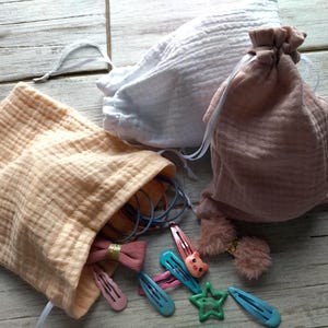 May include: Three muslin drawstring bags in peach, white, and mauve. The peach bag is open, revealing hair accessories like pink and blue barrettes, hair ties, and a star-shaped clip. The other bags are closed.