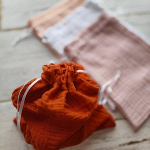 A pumpkin drawstring bag is in focus, with a textured fabric and white ribbon closure. Behind it, three other bags in shades of pink, white, and peach are slightly blurred. Perfect as aa vegetable storage bags.
