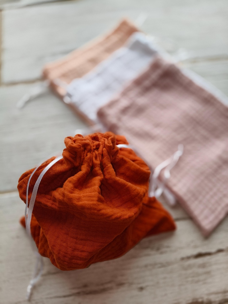 A pumpkin drawstring bag is in focus, with a textured fabric and white ribbon closure. Behind it, three other bags in shades of pink, white, and peach are slightly blurred. Perfect as aa vegetable storage bags.