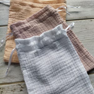 May include: Three muslin drawstring bags in shades of peach, mauve, and white. Each bag has a gathered top and a white satin ribbon drawstring. The bags are made of a textured, crinkled fabric.