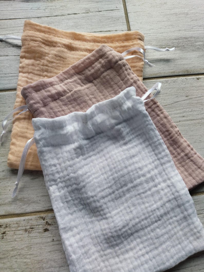 May include: Three muslin drawstring bags in shades of peach, mauve, and white. Each bag has a gathered top and a white satin ribbon drawstring. The bags are made of a textured, crinkled fabric.