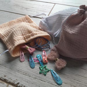 May include: Three muslin drawstring bags in peach, white, and pink, with various hair accessories. The peach bag is open, revealing hair ties and a pink bow clip. Several colorful snap clips and a green star-shaped clip are scattered around.