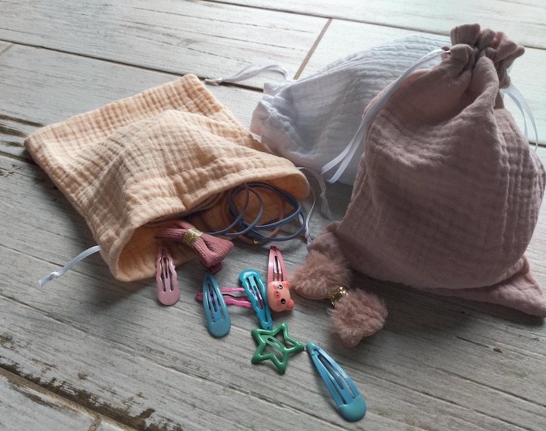 May include: Three muslin drawstring bags in peach, white, and pink, with various hair accessories. The peach bag is open, revealing hair ties and a pink bow clip. Several colorful snap clips and a green star-shaped clip are scattered around.