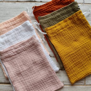 Puede incluir: Una colección de bolsas de muselina con cordón en varios colores, como melocotón, blanco, rosa, óxido, verde oliva y amarillo mostaza. Las bolsas, hechas de tela texturizada, están dispuestas sobre una superficie de madera clara.