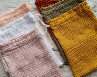 Puede incluir: Una colección de bolsas de muselina con cordón en varios colores, como melocotón, blanco, rosa, óxido, verde oliva y amarillo mostaza. Las bolsas, hechas de tela texturizada, están dispuestas sobre una superficie de madera clara.