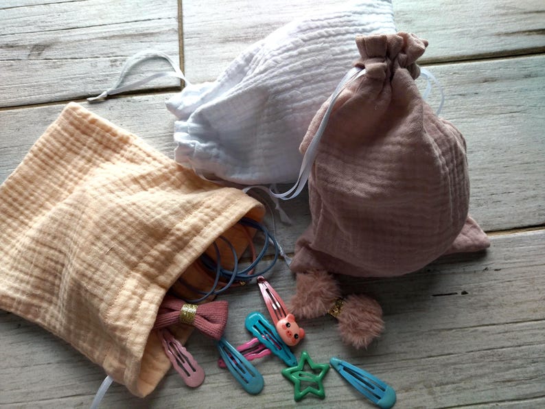 May include: Three muslin drawstring bags in peach, white, and mauve. The peach bag is open, revealing hair accessories like pink and blue barrettes, hair ties, and a star-shaped clip. The other bags are closed.