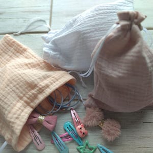 May include: A collection of hair accessories, including barrettes, bows, and scrunchies, are displayed alongside three drawstring pouches. The pouches are in shades of white and pink, made of a textured fabric. The accessories are in various colors, including pink, blue, and green.