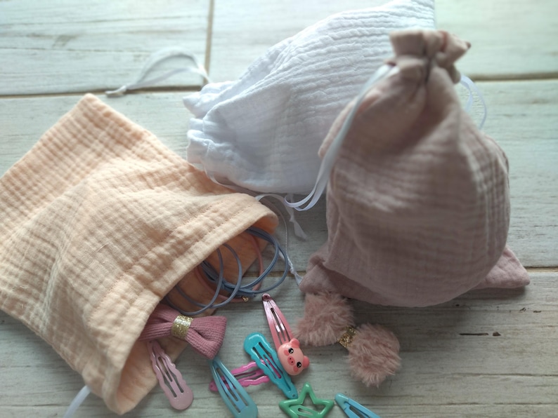 May include: A collection of hair accessories, including barrettes, bows, and scrunchies, are displayed alongside three drawstring pouches. The pouches are in shades of white and pink, made of a textured fabric. The accessories are in various colors, including pink, blue, and green.