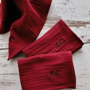 May include: Three deep red muslin cloths with a crinkled texture. One cloth features the embroidered letter C, while the other two cloths are each embroidered with the letters AC in a dark thread, providing a clear contrast against the fabric.