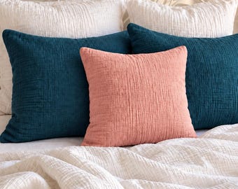May include: A collection of square throw pillows and a textured blanket. Two pillows are white, two are deep teal, and one is a soft coral pink. All items feature a crinkled, multi-layered fabric texture, arranged neatly on a bed with a metal headboard.