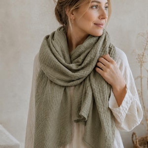 May include: A sage green scarf made of a lightweight, crinkled fabric with a soft, textured appearance. The scarf is draped loosely around the neck, featuring a rectangular shape with finished edges and a consistent, muted olive-green color throughout.