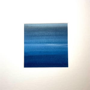 May include: A square watercolor painting featuring horizontal bands of blue hues, ranging from light to deep indigo. The artwork is set within a white mat, creating a clean, minimalist aesthetic. The painting evokes a sense of calm and depth.