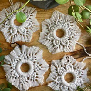 May include: Four white macrame star-shaped wall hangings with fringe details. The hangings are arranged on a wooden surface with a green plant in the background.