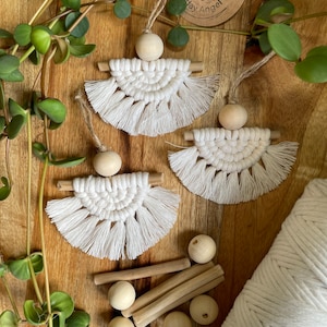 May include: Three white macrame angel ornaments with wooden beads and dowels, displayed on a wooden surface with greenery. The angels have a fringed design. Additional wooden beads, dowels, and a spool of white cord are also visible.