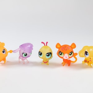 Puede incluir: Cinco figuritas de plástico de colores de animales pequeños. Los animales son una mariposa amarilla, un caniche morado, un gato naranja, un perro amarillo y un gato naranja claro. Las figuritas están todas en fila sobre un fondo blanco.