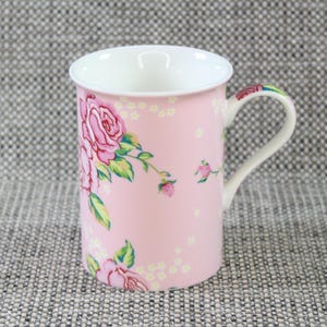 Può includere: Una tazza in ceramica rosa con un motivo floreale con rose rosa e foglie verdi. La tazza ha un manico bianco.