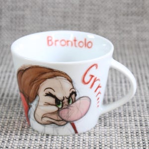 Puede incluir: Una taza de cerámica blanca con una ilustración colorida del personaje Gruñón con un gorro marrón y una nariz grande de color rosa. La palabra Brontolo está impresa en rojo en el borde interior y el texto rojo Grrr aparece en el exterior.